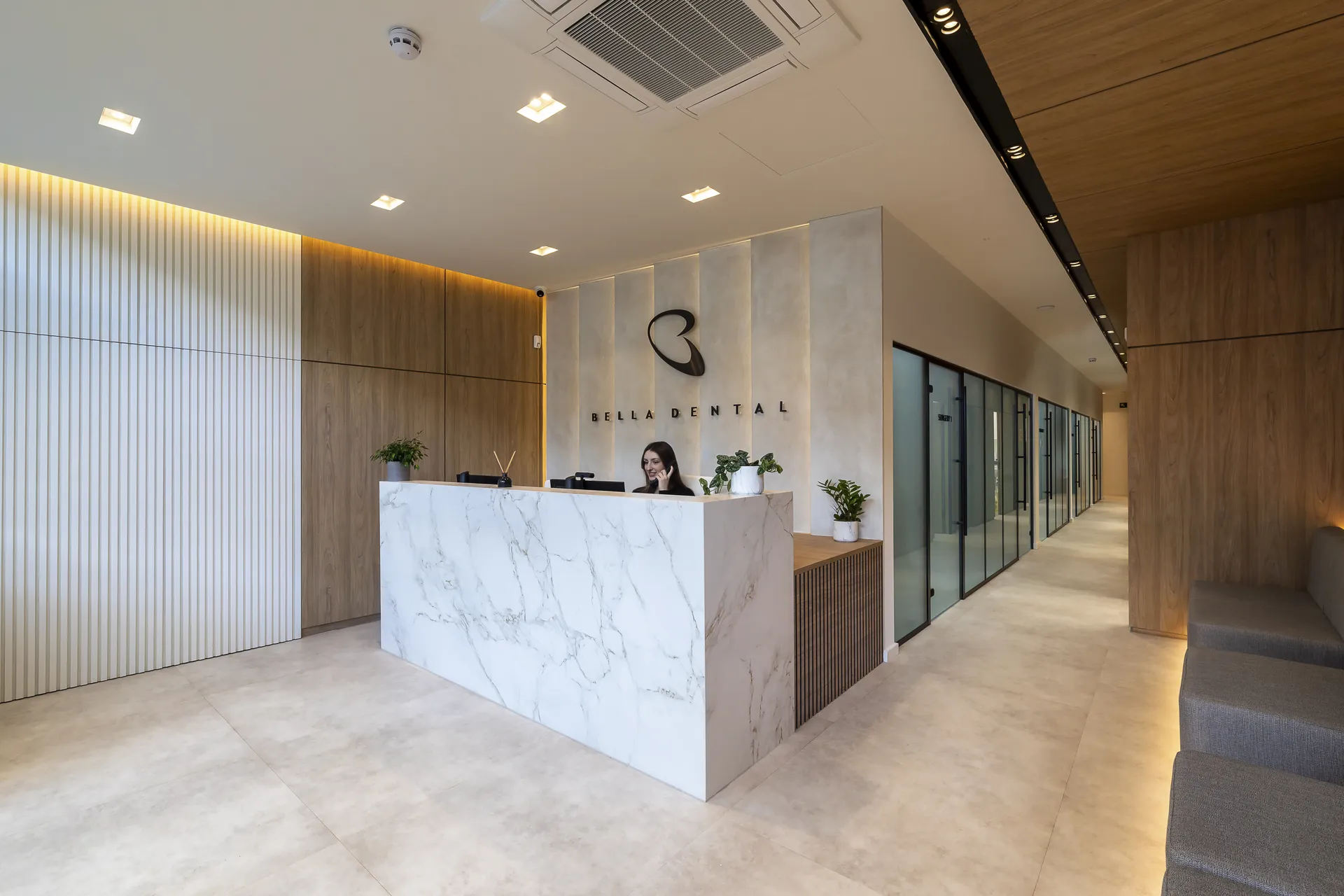 Modern dental clinic reception with a white marble counter, a receptionist on the phone, potted plants, a logo reading 'Bella Dental' on the wall, glass doors along a hallway, and seating with wooden paneling.