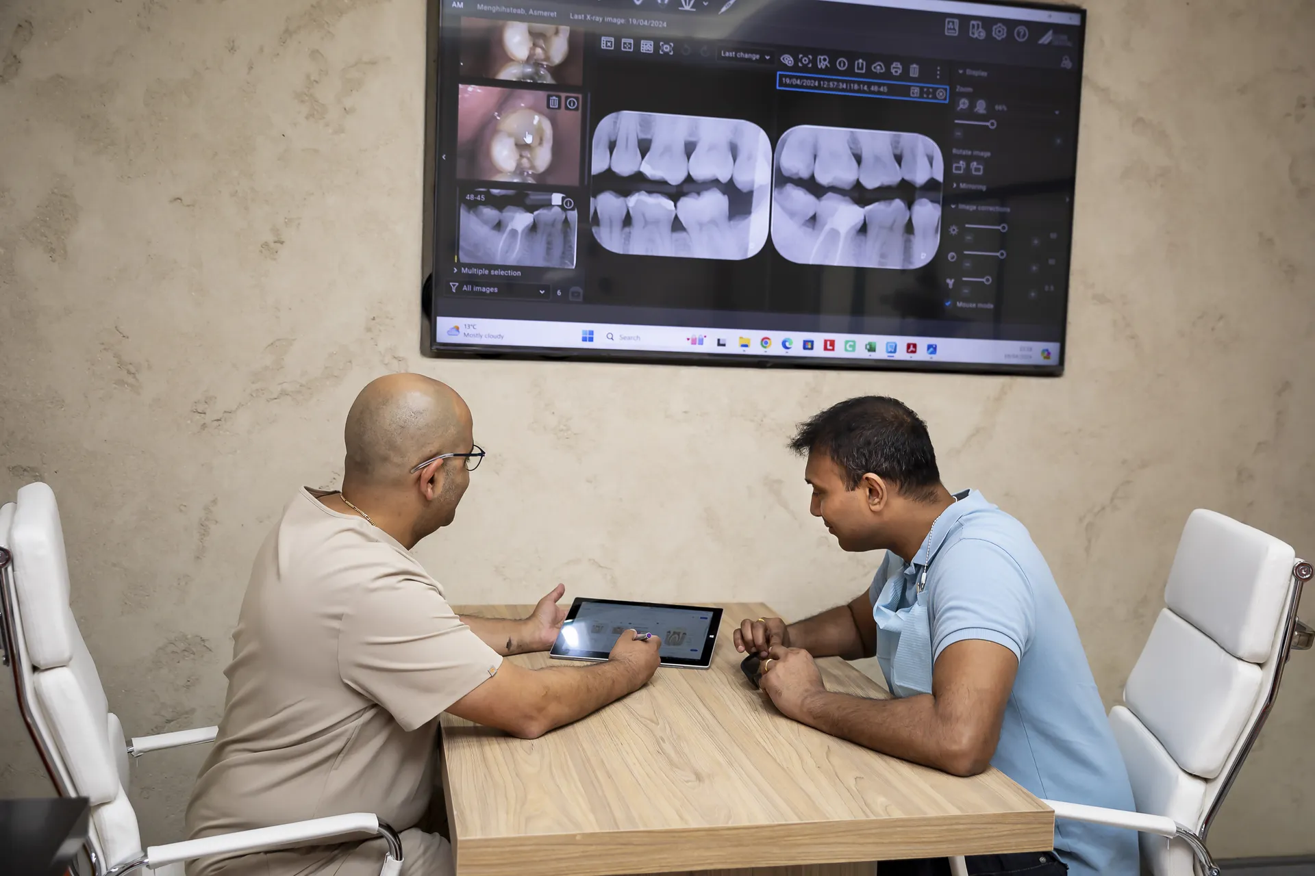 A dentist shows dental X-rays on a tablet to a male patient seated across a wooden table, with larger X-ray images displayed on a wall-mounted screen behind them.