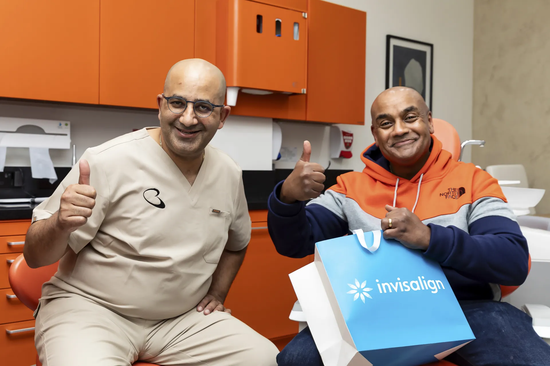 Two smiling men giving thumbs up in a dental office; one man is wearing beige scrubs, and the other in an orange and gray hoodie holds a blue Invisalign bag.