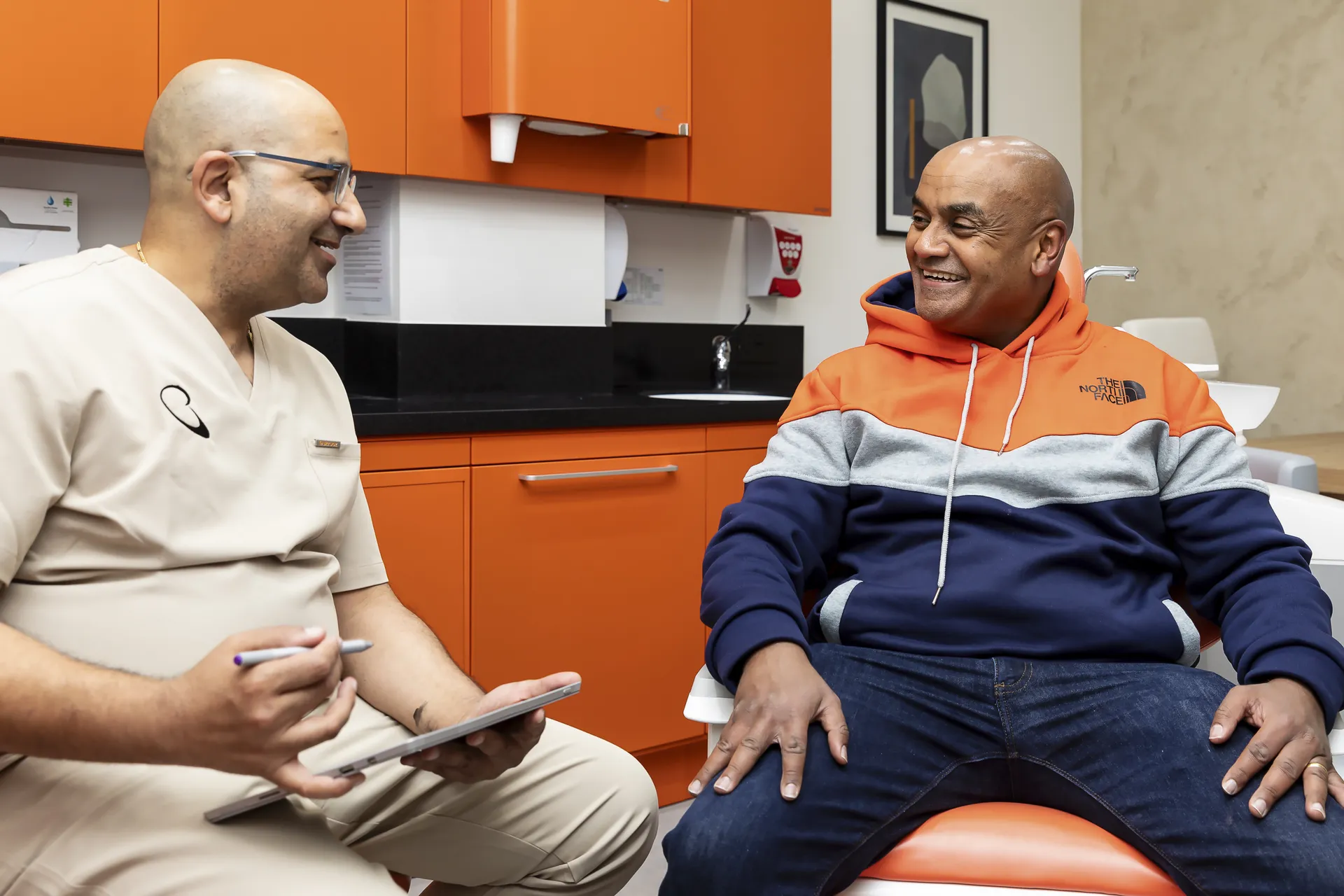 A healthcare professional in beige scrubs talking and smiling with a seated man wearing a blue, gray, and orange hoodie in a room with orange cabinets.