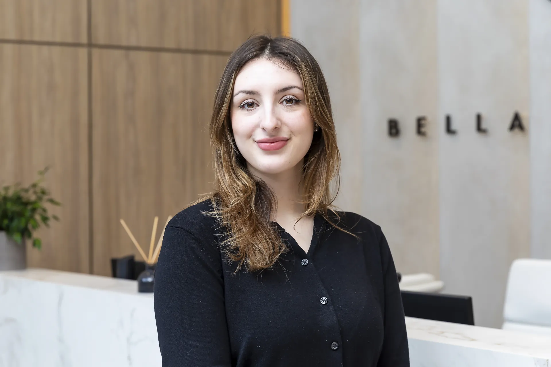 Young woman with light brown hair wearing a black buttoned cardigan, standing and smiling in a modern reception area with wooden panels and a marble counter, with the word BELLA partially visible on the wall behind her.