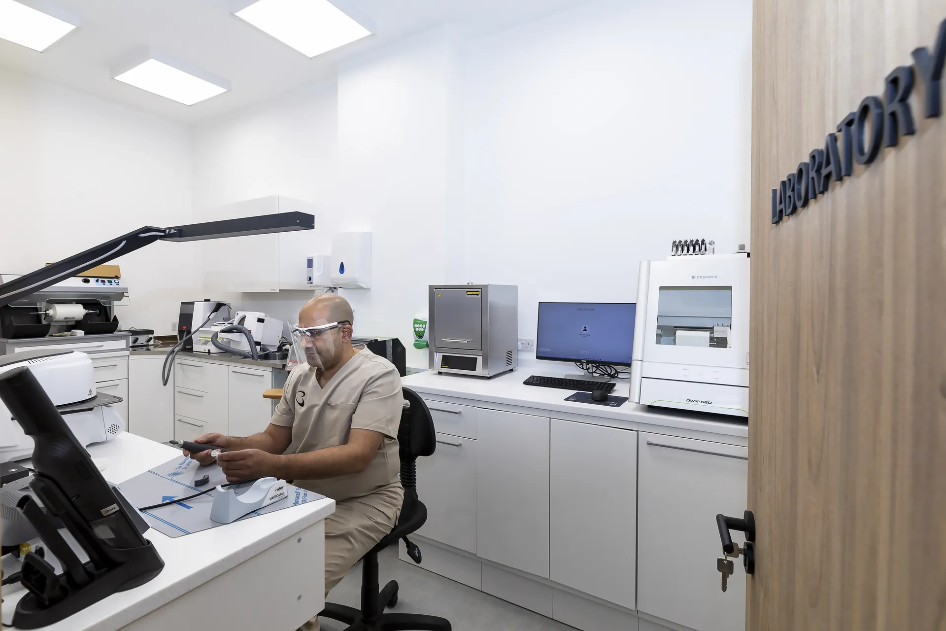 A man wearing beige scrubs, protective goggles, and a face shield works with dental or laboratory equipment inside a modern laboratory room with white cabinets and various machines, including a computer and sterilizer.