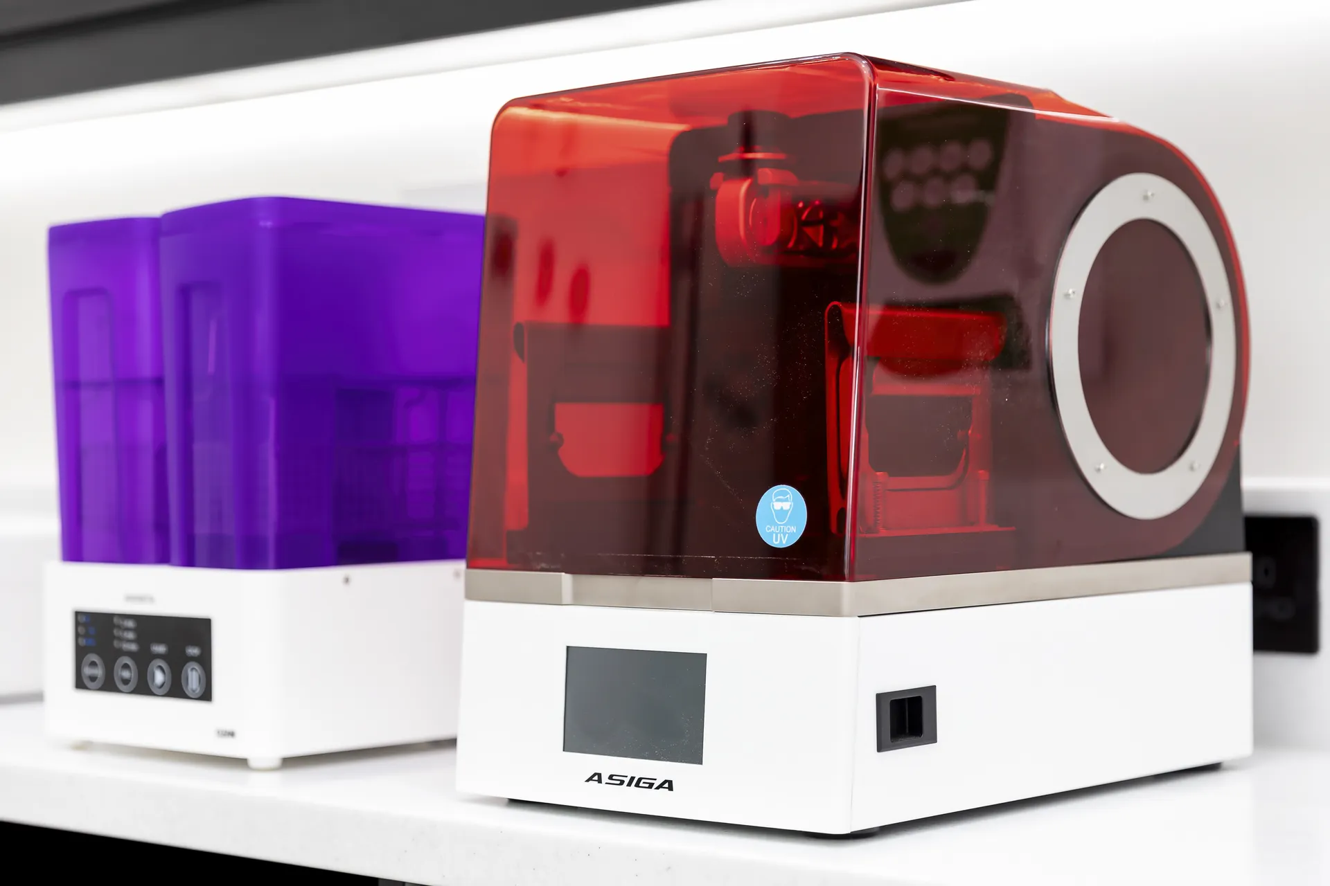 Two desktop 3D printers with translucent covers on a white countertop; the one in front has a red cover with a caution UV sticker and a display screen labeled ASIGA, and the one behind it has a purple cover with control buttons.