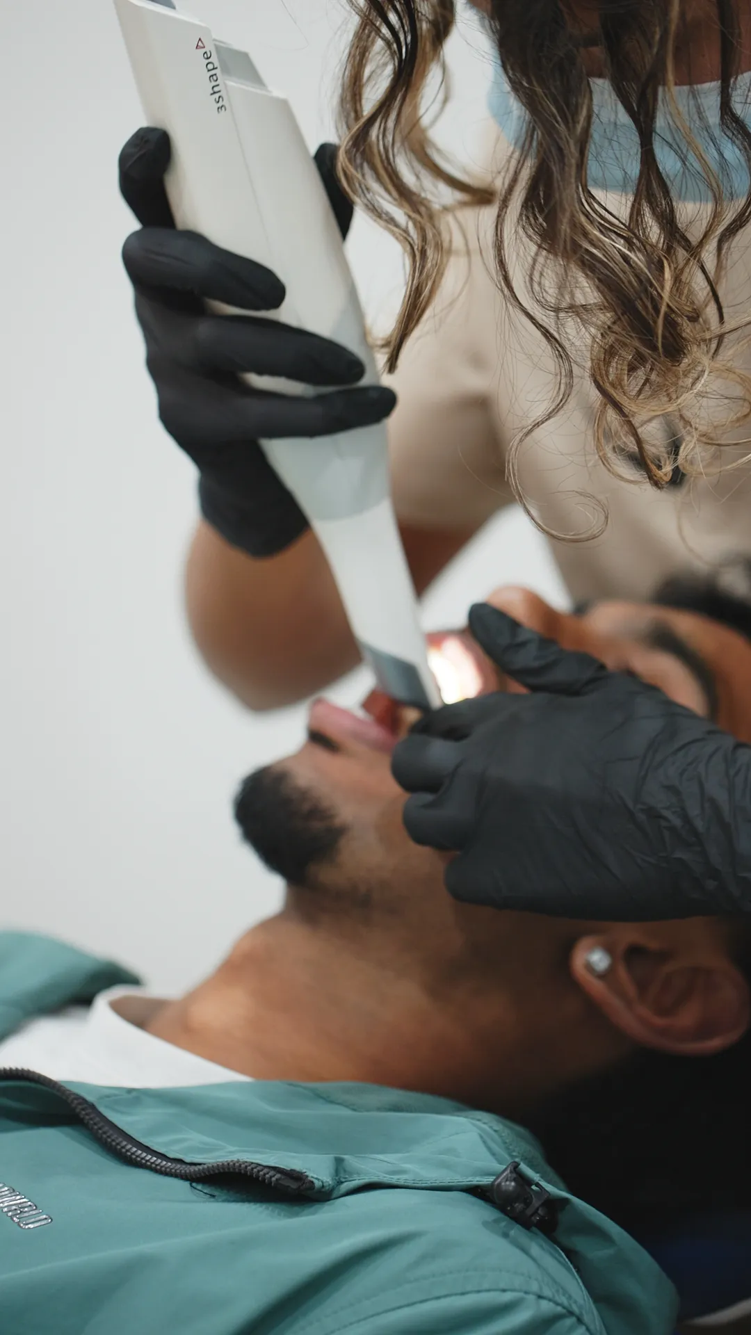 Close-up of a dental professional wearing black gloves using a 3shape intraoral scanner inside the mouth of a male patient reclining in a dental chair wearing a teal jacket and an earring.