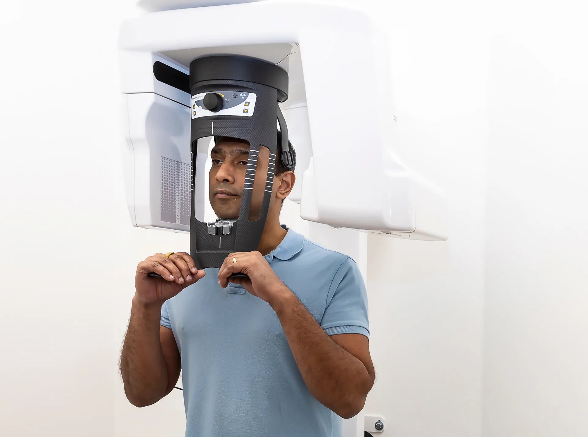 Man wearing a light blue polo shirt positioned in a dental panoramic X-ray machine, holding the black head support frame around his face.